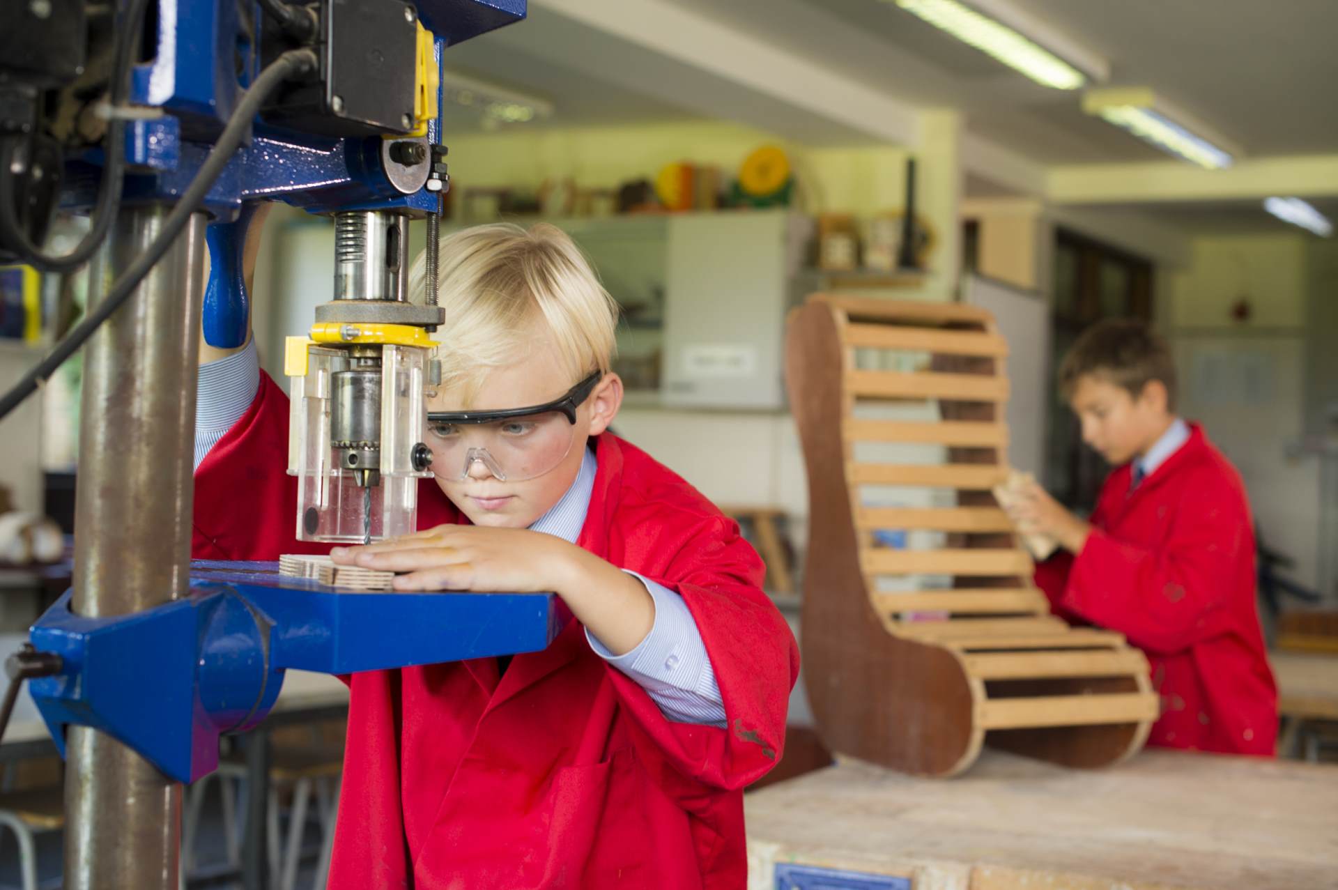 Two children in a design technology workshop. The child in the foreground, wearing safety glasses and a red coat, skillfully operates a drill press. The other child in the background diligently works on a wooden chair frame. Both are intensely focused on their creative tasks.