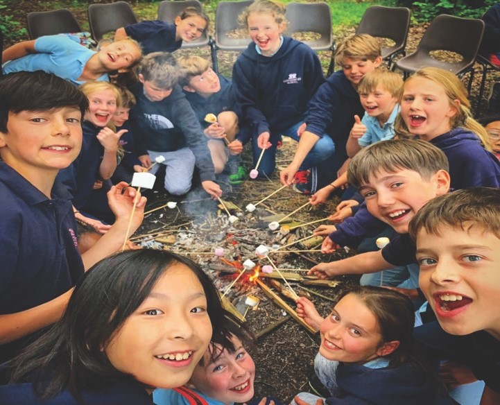A group of children gathered around a campfire, smiling as they roast marshmallows on sticks. Embracing the spirit of outdoor pursuits, they enjoy the camping adventure together with chairs visible in the background, soaking in every joyful moment.