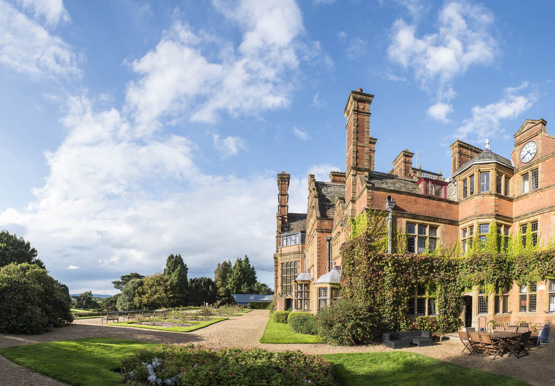 The School, a large historic brick building with ivy-covered walls, stands under a blue sky with scattered clouds. In the foreground, a gravel path and manicured garden lay amidst dense greenery surrounding the scene.