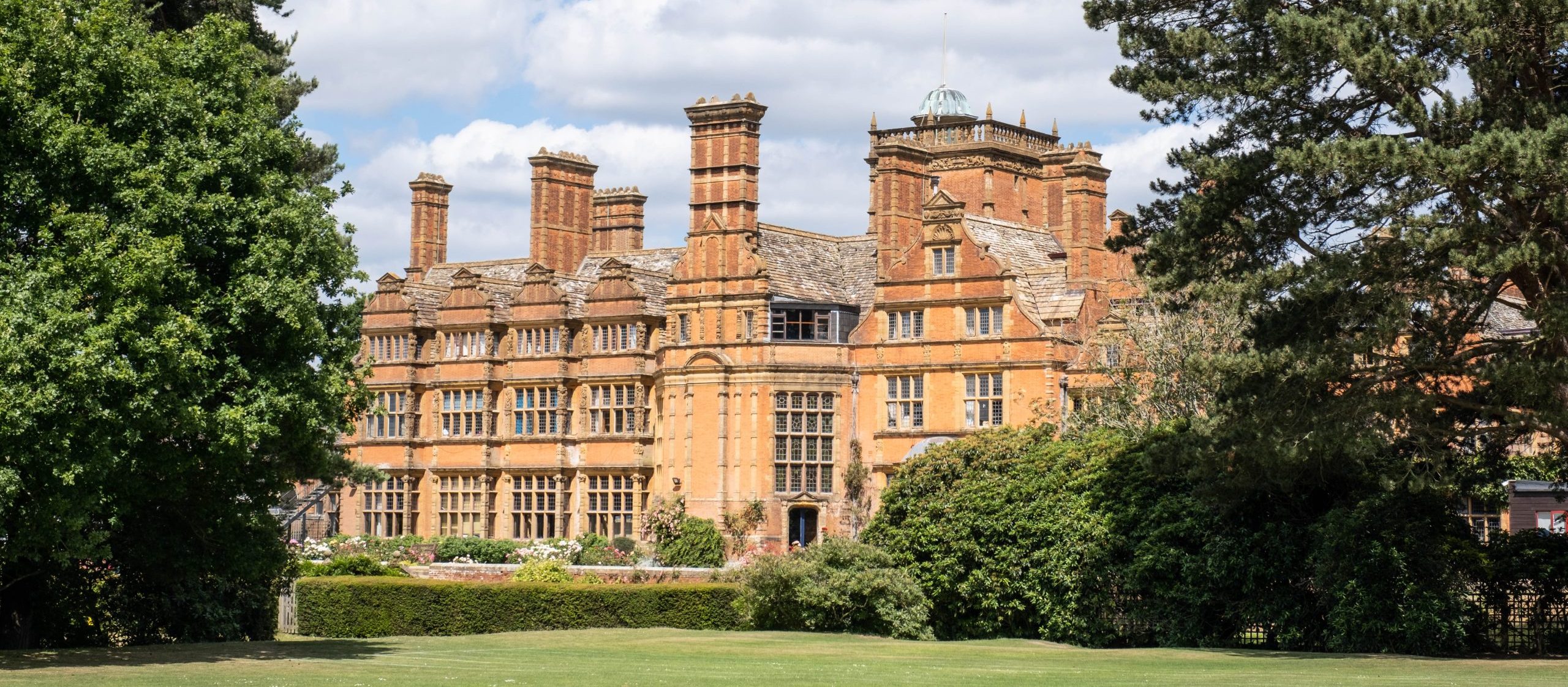 Historic brick mansion with multiple chimneys and an ornate roof, part of a prestigious institutions admissions tour, nestled amidst lush green trees and grass under a partly cloudy sky.