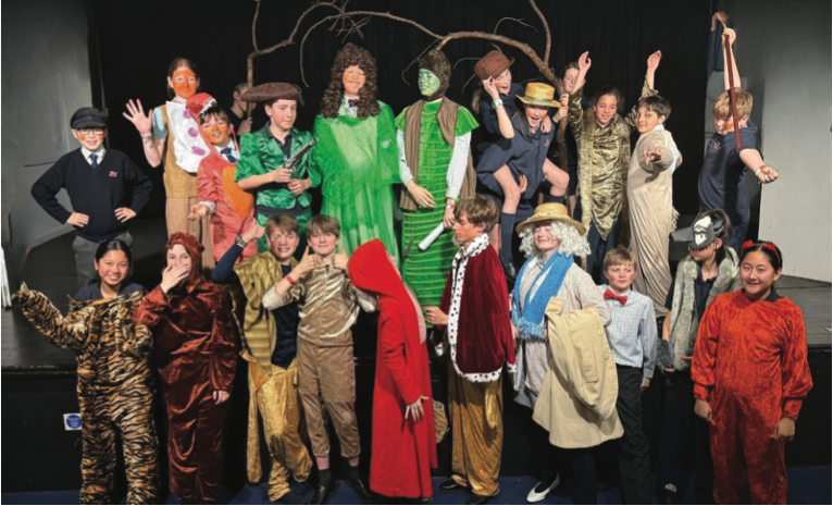 A group of children in colorful animal and fantasy costumes pose together on stage, smiling and making playful gestures, with a black backdrop and tree props behind them for the Newsletter - Michaelmas Term 2024.